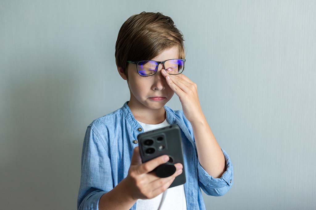 A young boy rubbing his eye holding smartphone.